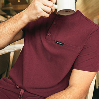 Man wearing burgundy color scrub top and scrub pant while drinking from a mug. - Men Nav Tile